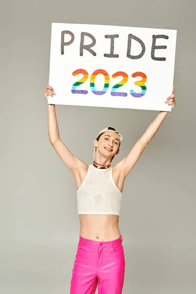 Tattooed and positive gay man in baseball cap, crop top, and pink pants holding pride 2023 placard above head while standing and looking at camera on grey background — Stock Photo