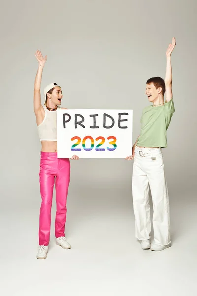 Happy lgbt friends in colorful clothes holding pride 2023 placard while raising hands and celebrating lgbtq community holiday in june on grey background in studio — Stock Photo