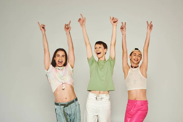 Positive and young lgbt community friends in colorful clothes smiling while looking at camera, raising hands and celebrating pride month together on grey background in studio — Stock Photo