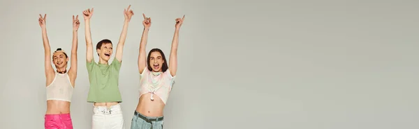 Happy and young lgbt community friends in colorful clothes smiling while looking at camera, raising hands and celebrating pride month together on grey background in studio, banner — Stock Photo