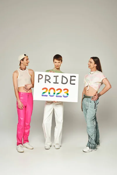 Happy gay men in colorful clothes standing and looking at queer friend holding pride 2023 placard while celebrating lgbt community holiday in June, grey background, studio — Stock Photo