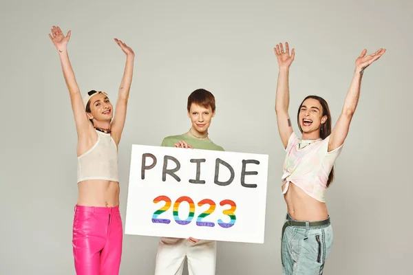 Happy and tattooed gay men in colorful clothes standing with raised hands near queer friend holding pride 2023 placard while celebrating lgbt community holiday in June, grey background, studio — Stock Photo