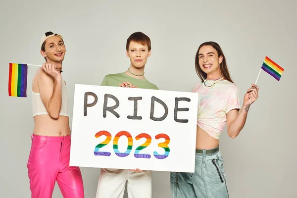 Happy and tattooed gay men in colorful clothes standing with rainbow flags near queer friend holding pride 2023 placard while celebrating lgbt community holiday in June, grey background, studio — Stock Photo