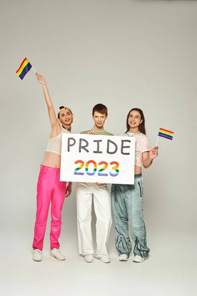 Cheerful and tattooed gay men in colorful clothes standing with rainbow flags near queer friend holding pride 2023 placard while celebrating lgbt community holiday, grey background, studio — Stock Photo