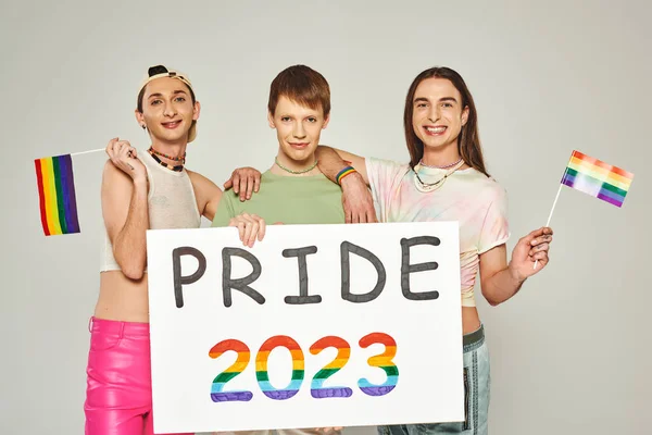 Positive and tattooed gay men standing with lgbt flags near queer friend holding pride 2023 placard while celebrating holiday in june, grey background, studio — Stock Photo