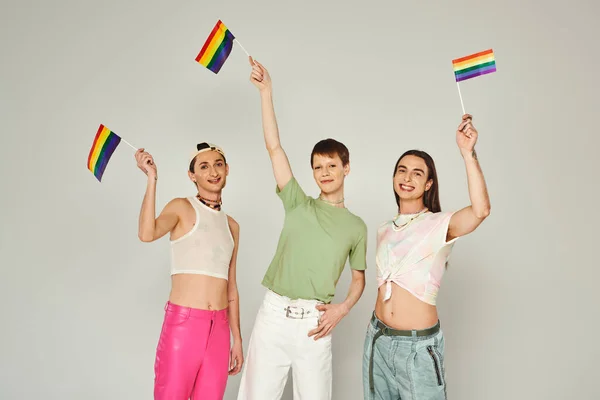 Happy and young lgbtq community friends with tattoos standing in colorful clothes and holding rainbow flags while looking at camera in studio, grey background — Stock Photo