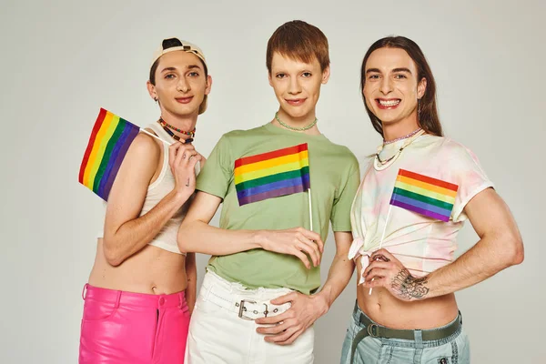 Positive and young lgbt friends with tattoos standing in colorful clothes and holding rainbow flags while looking at camera in studio on pride month — Stock Photo