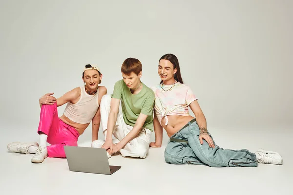 Diverso grupo de amigos positivos y jóvenes lgbt con tatuajes sentados juntos en ropa colorida y utilizando el ordenador portátil en el estudio sobre fondo gris durante el mes de orgullo - foto de stock