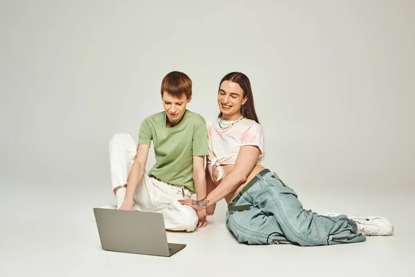 Positivo e jovem gay homem no colorido roupas sentado ao lado de tatuado amigo e usando laptop juntos no estúdio no cinza fundo durante orgulho mês — Fotografia de Stock