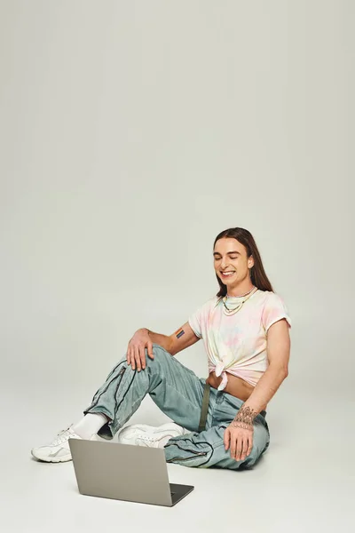 Positive and young gay man with long hair and trendy clothes sitting and looking at modern laptop in studio on grey background during pride month — Stock Photo