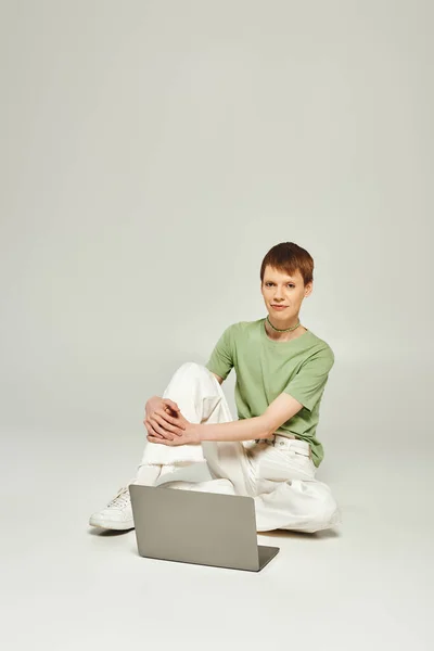Giovane uomo queer in t-shirt verde e jeans in denim bianco seduto e guardando la fotocamera vicino al computer portatile moderno in studio su sfondo grigio durante il mese dell'orgoglio — Foto stock