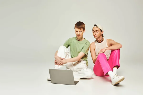 Young gay man in colorful clothes sitting next to friend in baseball cap and looking at laptop together in studio on grey background during pride month — Stock Photo