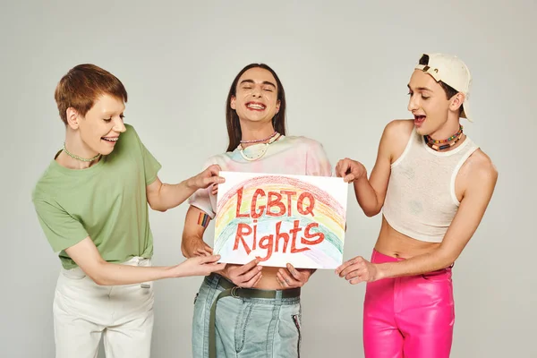 Happy lgbt friends in colorful clothes holding placard with lgbtq rights lettering while standing together and smiling on pride month, grey background — Stock Photo