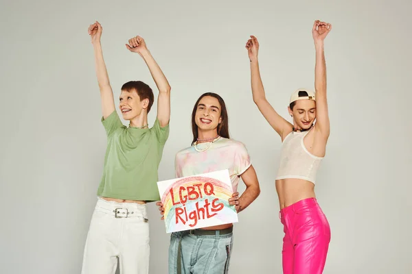 Щасливі друзі ЛГБТ в барвистому одязі танцюють з піднятими руками поруч з активістом, тримаючи плакат з правами ЛГБТкака і посміхаючись на місяць гордості, сірий фон — стокове фото