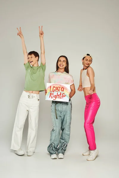 Happy lgbt friends in colorful clothes dancing next to positive gay activist holding placard with lgbtq rights lettering and smiling on pride month, grey background — Stock Photo