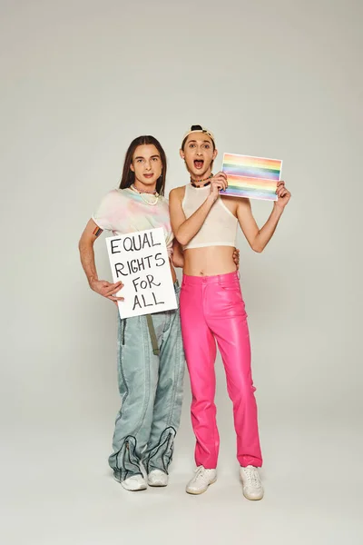 Emotional and tattooed lgbt people holding rainbow flag picture and placard with equal rights for all lettering while standing together on pride day, grey background — Stock Photo