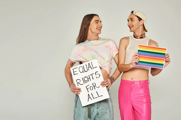 Positive and tattooed lgbt people holding rainbow flag picture and placard with equal rights for all lettering while looking at each other on pride day, grey background — Stock Photo