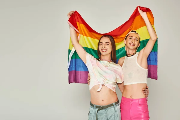 Alegre y tatuado gay personas en colorido ropa y desnudo vientre celebración arco iris lgbt bandera y abrazos mientras de pie juntos en orgullo día en gris fondo - foto de stock