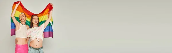 Allegri uomini gay in abiti colorati e ventre nudo con arcobaleno bandiera lgbt e abbracci mentre stanno insieme il giorno dell'orgoglio su sfondo grigio, banner — Foto stock