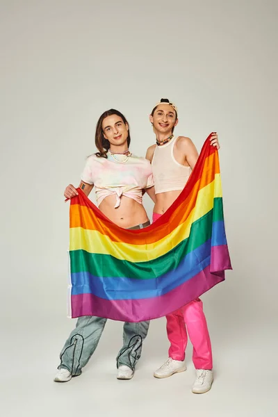 Cheerful and young lgbtq community friends in colorful clothes holding rainbow lgbt flag and looking at camera, standing together on pride day on grey background — Stock Photo