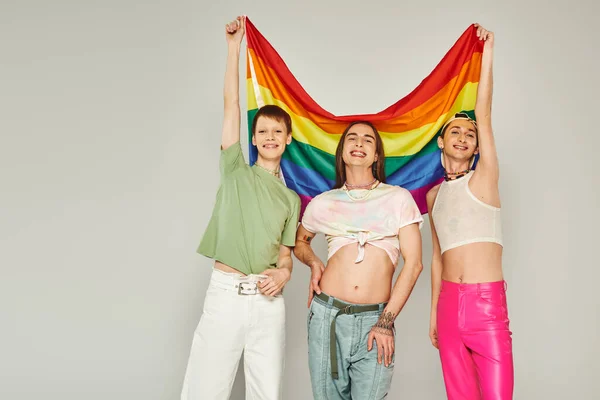 Divers groupe de gays joyeux en vêtements colorés regardant la caméra et tenant le drapeau lgbt arc-en-ciel tout en se tenant ensemble le jour de la fierté sur fond gris — Photo de stock