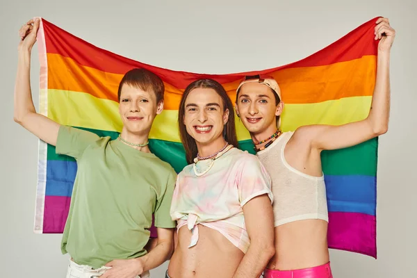 Diverso grupo de gente gay feliz en ropa colorida sosteniendo la bandera del arco iris lgbt y de pie juntos mientras se mira a la cámara en el día del orgullo sobre fondo gris - foto de stock