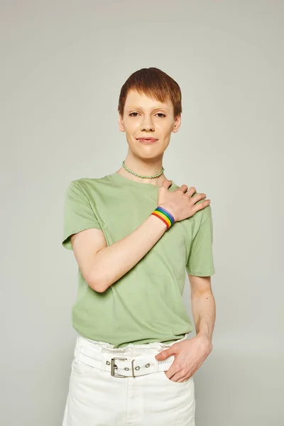 Porträt einer jungen queeren Person mit Lipgloss, die in grünem T-Shirt steht und die Hand mit lgbt-Fahnenarmband zeigt, während sie in die Kamera schaut, während der Stolz Monat auf grauem Hintergrund — Stockfoto