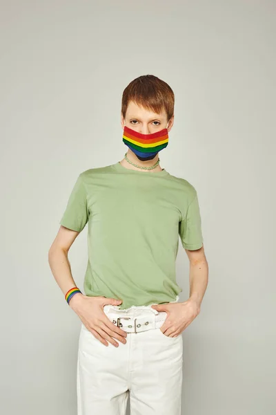 Портрет молодого странного человека, стоящего в зеленой футболке и маске с флагом lgbt, глядя в камеру и позируя во время месяца гордости на сером фоне — стоковое фото