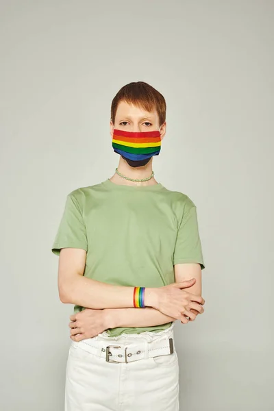 Retrato de jovem gay homem de pé em verde t-shirt e lgbt bandeira máscara enquanto olhando para câmera e posando durante o orgulho mês no fundo cinza — Fotografia de Stock