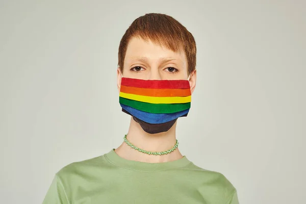 Portrait of young queer person standing in green t-shirt and lgbt flag mask while looking at camera during pride day on grey background — Stock Photo