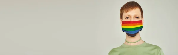 Портрет молодого странного человека, стоящего в зеленой футболке и маске с радужным флагом, смотрящего в камеру во время дня гордости на сером фоне, баннер — стоковое фото