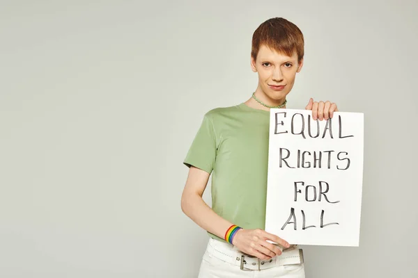 Portrait of young queer activist wearing lgbt flag bracelet and holding placard with equal rights for all lettering looking at camera during pride month on grey background — Stock Photo