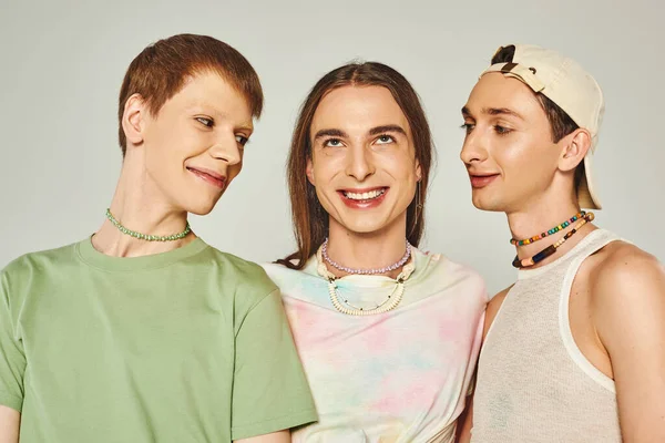Retrato de tres personas lgbtq con cuentas de colores sonriendo mientras están de pie juntos sobre fondo gris en el estudio, celebración del concepto del mes de orgullo - foto de stock