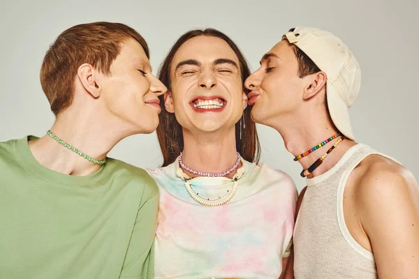 Portrait of happy gay men with colorful beads smiling while kissing cheeks of friend on grey background in studio, celebration of pride month concept — стоковое фото