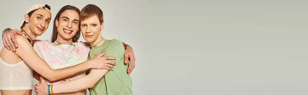 Retrato de três pessoas lgbtq com contas coloridas sorrindo ao olhar para a câmera e abraçando uns aos outros em fundo cinza no estúdio, celebração do conceito de mês de orgulho, banner — Fotografia de Stock