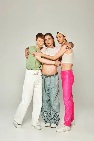 Полный рост трех lgbtq людей в цветастых одеждах, смотрящих на камеру и обнимающих друг друга на сером фоне в студии, празднование концепции месяца гордости — стоковое фото