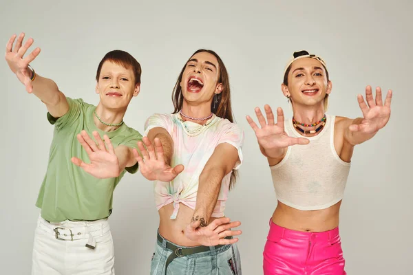 Cheerful lgbtq friends in colorful clothes looking at camera and standing with outstretched hands on grey background in studio, celebration of pride month concept — Stock Photo
