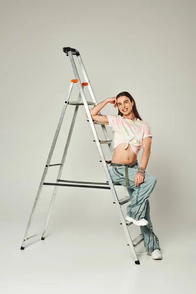 Татуированный и веселый гей с длинными волосами, позирующий в джинсах в джинсах с голым животом, сидящий на лестнице во время месяца гордости на сером фоне — стоковое фото