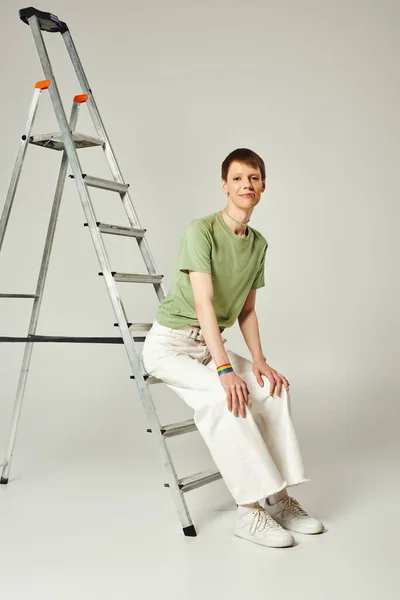 Full length of happy queer person with shiny lip gloss posing in green t-shirt and white denim jeans while sitting on ladder during pride month on grey background — Stock Photo