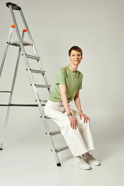 Personne queer heureux pleine longueur avec brillant à lèvres brillant posant en t-shirt vert et jeans en denim blanc tout en étant assis sur l'échelle pendant le mois de la fierté sur fond gris — Photo de stock