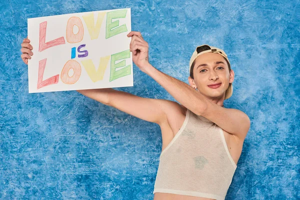 Cheerful young gay activist in baseball cap and white tank top smiling while holding placard with love is love words during pride month on mottled blue background — Stock Photo