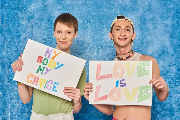 Activistas gays positivos sonriendo mientras sostienen pancartas con amor es amor y mi cuerpo mis palabras de elección durante el mes de orgullo sobre fondo azul moteado - foto de stock