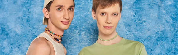 Young lgbtq friends with colorful beads and casual clothes looking at camera while standing together on mottled blue background during pride month, banner — Stock Photo