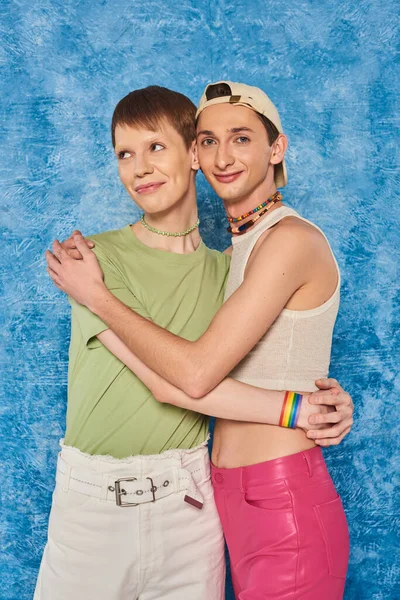 Молодые друзья lgbtq с красочными бусами, стоящими в повседневной одежде и обнимающими друг друга, улыбаясь на пятнистом голубом фоне во время месяца гордости — стоковое фото