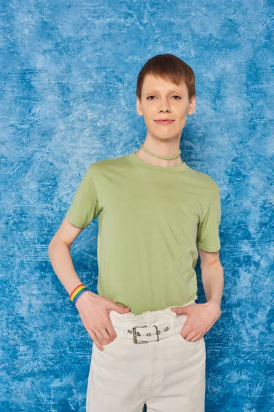 Jeune queer en vêtements décontractés tenant la main dans des poches de pantalon et regardant la caméra pendant la célébration du mois de fierté lgbt sur fond bleu moucheté — Photo de stock
