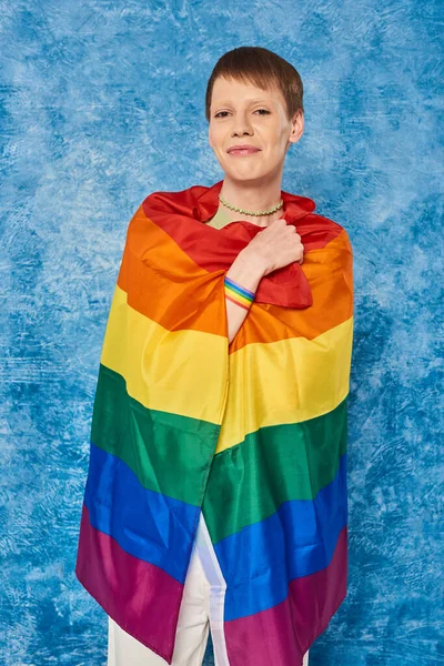 Retrato de alegre persona queer sonriendo mientras sostiene la bandera lgbt y mirando a la cámara durante la celebración del mes del orgullo gay sobre fondo azul moteado - foto de stock