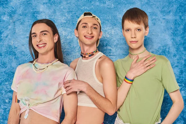 Веселые друзья-гомосексуалисты в повседневной одежде смотрят в камеру и празднуют месяц гордости, стоя вместе на пестром синем фоне — стоковое фото