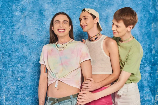 Jóvenes amigos queer mirando a amigo gay en ropa casual mirando a la cámara durante lgbt orgullo mes celebración en textura moteado fondo azul - foto de stock