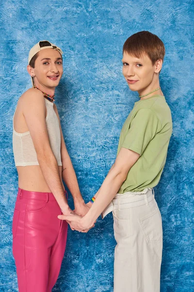 Sonrientes amigos homosexuales tomados de la mano y mirando a la cámara durante la celebración del mes del orgullo gay en un fondo azul texturizado y moteado - foto de stock