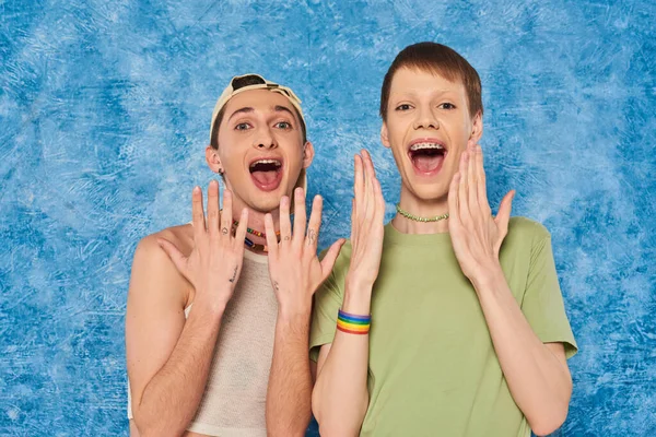 Шокированные друзья-гомосексуалисты в повседневной одежде открывают рот и смотрят в камеру во время празднования месяца гордости на пестром синем фоне — стоковое фото